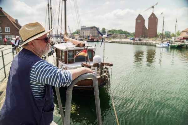 Axel Prahl am Hafen. Axel Prahl steht am Hafen und blickt auf das Wasser. Er trägt Sonnenbrille und Sonnenhut.