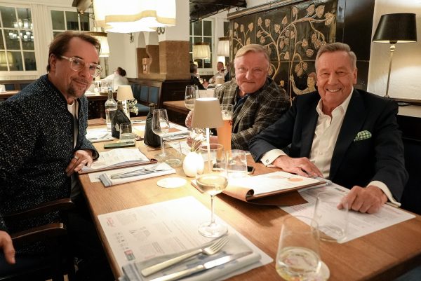 Jan Josef Liefers, Axel Prahl und Roland Kaiser©NDR WDR 2025 Jan Josef Liefers, Axel Prahl und Roland Kaiser sitzen am Tisch in einem Restaurant und lächeln in die Kamera.